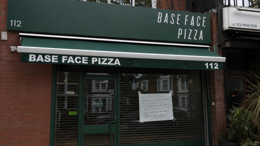 Handwritten basement flood notice displayed behind the security grille at Base Face Pizza in Richmond