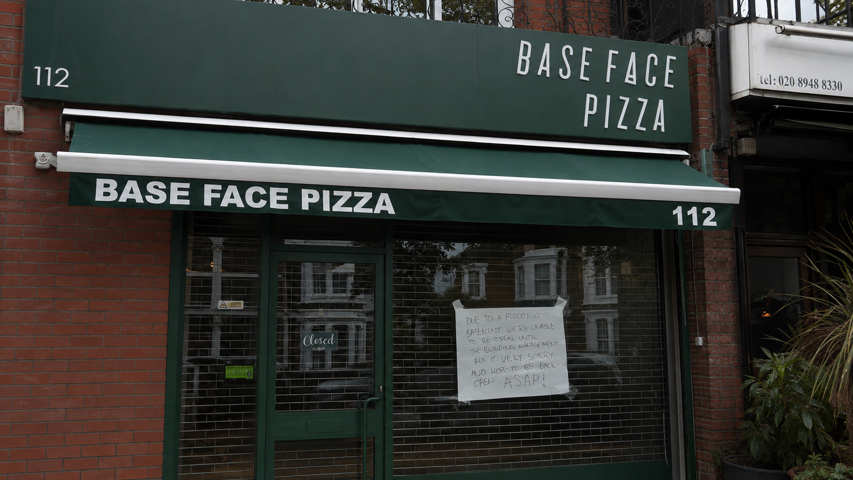 Handwritten basement flood notice displayed behind the security grille at Base Face Pizza in Richmond